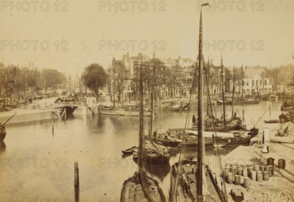 The Spanish Quay, View of the Spaansekade in Rotterdam, Rotterdam, cabinet photograph, annotation: '45, 5, 40, 12x: 15, 55', publisher: Andries Jager, Amsterdam, 1860 - 1890, photographer, paper, pasteboard, albumen print, height 108 mm x width 168 mm, canals, waters (in city), Spaansekade, A sepia-toned waterfront scene showing a quay lined with moored sailing vessels with tall masts and rigging, small rowboats alongside larger wooden boats, and stacks of barrels and crates on the dock; a stone embankment and a bridge span the waterway, buildings and trees rise behind the quay, and faint reflections of boats appear on the water surface.