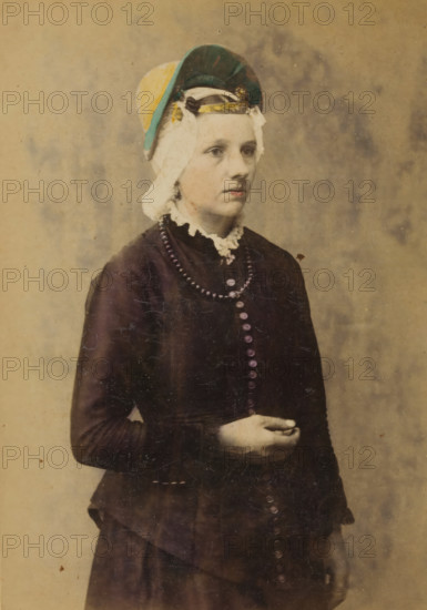 Portrait of an unknown woman with a headdress in the traditional costume of Zaandam, North Holland, La Hollande du Nord. Zaandam, Costumes of the Netherlands, cabinet photograph, inscription: 'Published by A. JAGER, Damrak 81, Amsterdam., Reproduction rights reserved.', retailer's mark: 'Boek-, Papier- en Kunsthandel., Founded 1813, A. VAN HOOGSTRATEN & Zn., Royal Warrant Holders, PLACE 11, - The Hague.', photographer: Andries Jager (possibly), publisher: Andries Jager, Amsterdam, 1860 - 1890, Andries Jager, photographer, publisher, paper, pasteboard, albumen print, height 168 mm x width 108 mm, folk costume, regional costume, historical persons not known by name - BB - woman, A young person shown from head to mid-thigh stands against a plain, textured backdrop. They wear a dark long-sleeved dress with a row of round buttons down the front and a high lace collar. A beaded necklace lies over the bodice. Their hair is covered by a layered headpiece featuring a light cloth, a darker round...