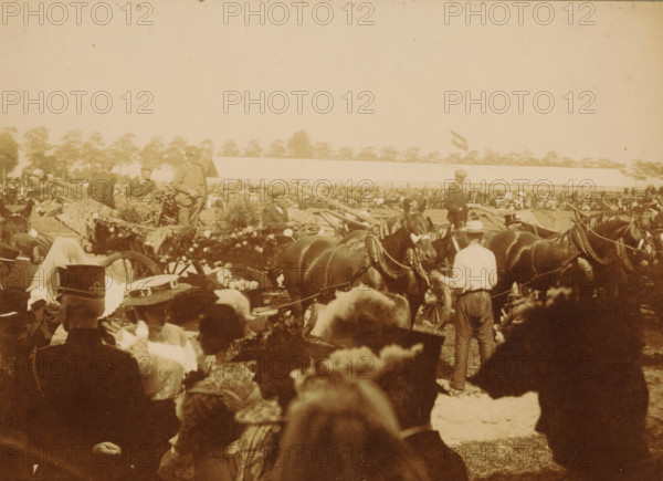 Show with horses, presumably in the Netherlands, photograph, Netherlands, c. 1895 - c. 1905, photographer, paper, pasteboard, baryta paper, daylight gelatin silver print, height 139 mm x width 177 mm, horse, A sepia-toned outdoor scene showing a crowd of people and a line of horses and decorated wagons. Several men in hats stand near the horses, some holding reins, while others stand on the wagons among bundles of hay and floral decorations. In the foreground, women wearing hats with ribbons and feathers are visible from behind and in profile. A long low building and a distant row of trees form the background, and a flag flies above the building. The overall image has a faded, vintage appearance.