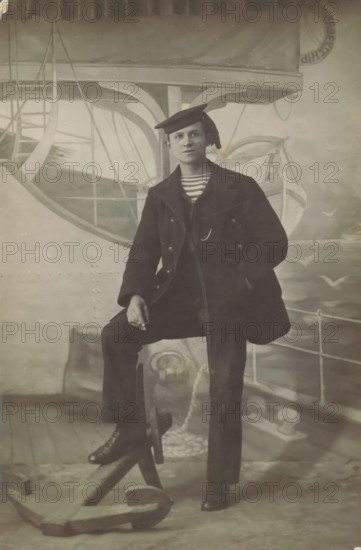 Studio portrait of an unknown man in sailor uniform, photograph, picture postcard, inscription: 'Chef Kreuger Koningstraat 88 Photographer, Den Helder', photographer: Jozeph Kreuger, Den Helder, c. 1915 - c. 1925, paper, gelatin silver print, developing gelatin silver print, height 138 mm x width 90 mm, seaman, studio, A seated person wearing a dark double-breasted coat, striped shirt visible at the collar, dark trousers, and a tilted flat cap sits with one leg raised on a wooden support and one hand resting on the raised knee; the other hand is in a pocket. The backdrop shows painted nautical elements including a lifeboat, rigging, railings, and a large anchor on the floor in front. The image is monochrome with soft lighting and visible surface scratches and creases.