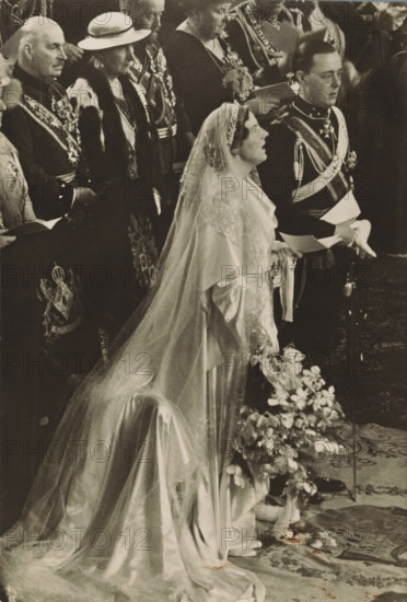 Wedding ceremony of Princess Juliana and Prince Bernhard in the St. Jacob's Church in The Hague on January 7, 1937, photograph, postcard, photographer: Erich Salomon, The Hague, publisher: Het Leven, Weekblad, Netherlands, 1937-01-07, Het Leven, Erich Salomon, photographer, printer (person), publisher, paper, gelatin silver print, height 149 mm x width 101 mm, Person, Juliana of the Netherlands, Bernhard (Prince of the Netherlands), (civil) marriage ceremonies, prince, crown princess, Wedding of Princess Juliana and Prince Bernhard, Grote Kerk (The Hague), A black and white photograph showing a bride in a long, flowing gown with a sheer veil and ornate headpiece standing beside a man in a formal uniform with decorative sashes and epaulettes; the bride holds a bouquet of flowers and her gown’s train spreads across a patterned floor covering, while behind them several seated and standing figures wear formal attire and regalia, including a woman with a hat and fur-trimmed coat and men wit...