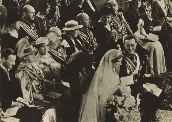 Wedding ceremony of Princess Juliana and Prince Bernhard in the St. Jacob's Church in The Hague on January 7, 1937, photograph, postcard, photographer: Erich Salomon, The Hague, publisher: Het Leven, Weekblad, 1937-01-07, Het Leven, Erich Salomon, photographer, printer (person), publisher, paper, gelatin silver print, height 104 mm x width 146 mm, Person, Juliana of the Netherlands, Bernhard (Prince of the Netherlands), (civil) marriage ceremonies, prince, crown princess, Wedding of Princess Juliana and Prince Bernhard, Grote Kerk (The Hague), A black-and-white photograph showing seated people dressed in formal attire and ceremonial uniforms, many wearing sashes, medals, chains, and hats; a woman in a veil and gown is in the foreground holding flowers, a man beside her in a decorated uniform is smiling, and several other adults in hats, robes, and fur-trimmed coats are seated behind them, some holding booklets.