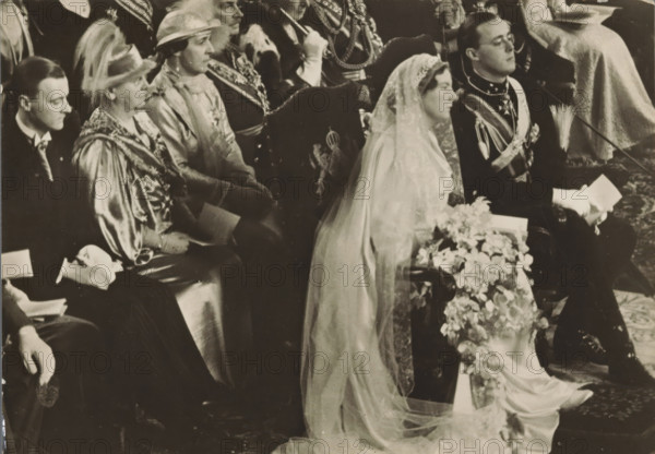 Wedding ceremony of Princess Juliana and Prince Bernhard in the St. Jacob's Church in The Hague on January 7, 1937, photograph, postcard, photographer: Erich Salomon, The Hague, publisher: Het Leven, Weekblad, 1937-01-07, Het Leven, Erich Salomon, photographer, printer (person), publisher, paper, gelatin silver print, height 104 mm x width 146 mm, Person, Juliana of the Netherlands, Bernhard (Prince of the Netherlands), (civil) marriage ceremonies, prince, crown princess, Marriage of Princess Juliana and Prince Bernhard, Flushing, Grote or Sint-Jacobskerk (Vlissingen), A black and white photograph showing a row of seated people dressed in formal attire, including a woman in a bridal gown with a long lace veil draped over her head and shoulders, holding a floral bouquet that cascades forward, and a man next to her in a dark uniform with decorative sashes and gloves holding a folded item. Other seated figures wear hats, ornate robes, and ceremonial garments, and some hold small booklets....