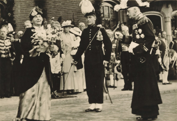 40th anniversary of the reign of HM the Queen, Amsterdam, 5 September 1938, arrival at Haarlemmermeer Station, photograph, postcard, Haarlemmermeer, publisher: Have, W. ten, 1938-09-05, photographer, printer (person), paper, gelatin silver print, height 104 mm x width 149 mm, Person, Wilhelmina of the Netherlands, queen, A black and white scene of formally dressed people gathered outdoors in front of a brick building, with a woman in a hat holding a large bouquet in the left foreground and a man in a dark uniform with a plumed hat standing center holding a sword, while another man in a dark ceremonial coat with ornate decorations and a feathered hat stands to the right; behind them are several onlookers in hats and suits, decorative sashes and uniforms, and architectural details including an arched doorway.