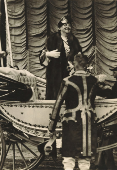40th anniversary of the reign of HM the Queen, Amsterdam, 5 September 1938, HM the Queen, upon arrival at Haarlemmermeer Station, photograph, postcard, publisher: Have, W. ten, 1938-09-05, photographer, printer (person), paper, gelatin silver print, height 149 mm x width 105 mm, Person, Wilhelmina of the Netherlands, queen, A sepia-toned scene showing a person standing in a decorative carriage wearing a dark fur-trimmed coat and a small hat, smiling and gesturing with one hand while a person with their back to the camera stands in front of the carriage wearing a patterned coat with vertical and diagonal stripes and light-colored footwear; the carriage features tufted upholstery and ornate trim, with visible wooden wheels and a curved side panel, and a draped, scalloped fabric backdrop fills the upper background.