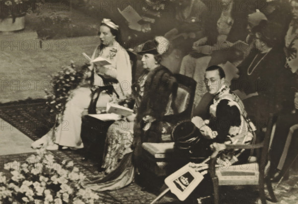 Nieuwe Kerk, Amsterdam, 6 September 1938. Commemorative service on the occasion of the 40th anniversary of the reign of HM the Queen, photograph, postcard, publisher: Have, W. ten, 1938-09-06, photographer, printer (person), paper, gelatin silver print, height 105 mm x width 149 mm, Person, Juliana of the Netherlands, Wilhelmina of the Netherlands, Bernhard (Prince of the Netherlands), queen, prince, crown princess, A sepia-toned scene shows a group of seated people dressed in formal, historic attire, with a floral arrangement in the foreground and patterned rugs beneath them. The central figures include a woman wearing a dark fur-trimmed coat and a hat with a decorative plume, seated next to a man in ornate clothing holding a round hat on his lap; both hold open booklets. To the left, another woman in a light dress and headpiece also holds an open booklet. Behind and around them, other attendees are visible, some holding papers, and a woman wearing a dark dress with a long necklace is...