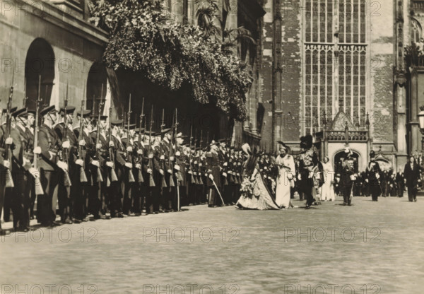40th anniversary of the reign of HM the Queen, Amsterdam, 6 September 1938. Return from the Nieuwe Kerk, photograph, postcard, publisher: Have, W. ten, 1938-09-06, photographer, printer (person), paper, gelatin silver print, gelatin silver developed, height 105 mm x width 149 mm, Person, Wilhelmina of the Netherlands, queen, Nieuwe Kerk (Amsterdam), A long line of uniformed soldiers stands in formation along the left side of a paved courtyard, each holding a rifle with an upright bayonet and wearing peaked caps and long coats with visible gloves; to the right of them a procession of formally dressed figures moves past, including a central woman in an ornate gown with a long train and floral adornments accompanied by other attendants in light-colored dresses and ceremonial attire, while uniformed officers in decorated uniforms walk nearby; the scene is framed by a large stone building with tall arched windows and a facade of brick and stone, and a dense canopy of foliage overhangs part ...