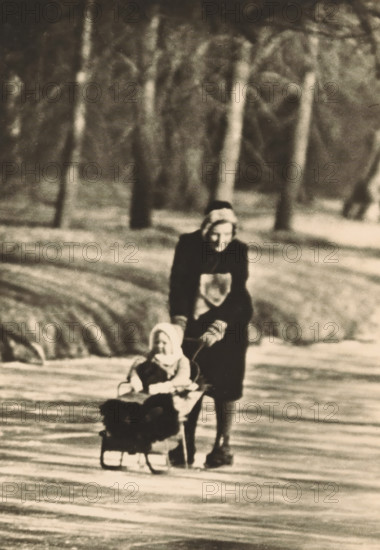 On the ponds of Huis ten Bosch, photograph, postcard, The Hague, publisher: Have, W. ten, Amsterdam, 1940-01-11, photographer, printer (person), paper, gelatin silver print, height 150 mm x width 104 mm, Person, Beatrix (Queen of the Netherlands), Juliana of the Netherlands, mother and daughter(s) (family group), sleigh, sledge (non-sport), princess, crown princess, Huis ten Bosch Palace, A sepia-toned scene showing a person wearing a dark coat and head covering pushing a small child seated in a sled across a snowy surface, with blurred vertical tree trunks and soft-focus snow in the background.