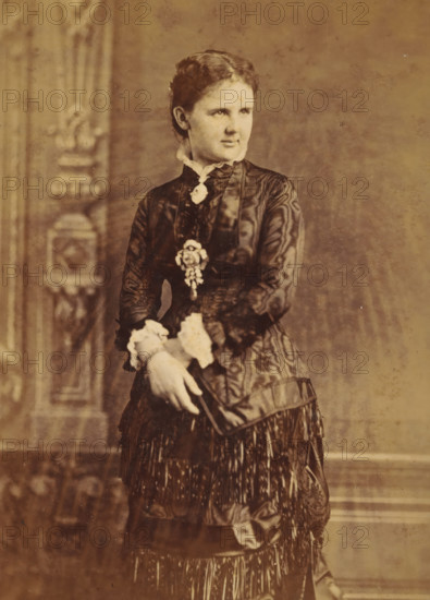 Portrait of Queen Emma, cabinet photograph, inscription: 'ML VERVEER PHOTOGRAPHE, DE LLMM LE ROI ET, LA REINE DES PAYS BAS, 32. Zeestraat, vis du Willemspark la Haye.', photographer: Maurits Verveer, The Hague, 1878 - 1890, pasteboard, paper, albumen print, albumendruk, height 166 mm x width 108 mm, Person, Emma (Queen consort of the Netherlands), historical persons, princess, A sepia-toned portrait of a young woman standing against a studio backdrop with ornate vertical patterns on the left and a plain textured surface on the right, wearing a dark satin dress with a high collar and decorative trim, lace cuffs visible at her wrists, a floral brooch pinned near her chest and a second ornamental piece attached lower on her bodice, her hair pulled back and styled with gentle waves, her hands clasping a thin object held near her skirt which features layered fabric and long fringe.