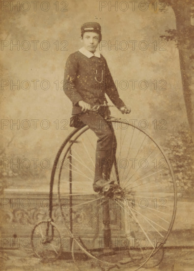 Portrait of Matthieu van Oosterzee on a bicycle, cabinet photograph, inscription: 'A. Lüssow Utrecht Zonstraat', photographer: Gustav Albert Fredrich Christoph Lüssow, Utrecht, c. 1855 - c. 1860, paper, pasteboard, albumen print, height 139 mm x width 98 mm, Person, Mattheus Jacobus van Oosterzee, boy (child between toddler and youth), bicycle, A sepia-toned photograph shows a young person seated on a high-wheeled bicycle, wearing a dark jacket with a white collar and a small cap; one hand rests on the handlebars and one foot is on a pedal, while the large front wheel and smaller rear wheel of the bicycle are prominent, with visible spokes and a curved metal frame, and the background features a mottled textured surface with faint vertical elements and a tree-like shape at the right edge.