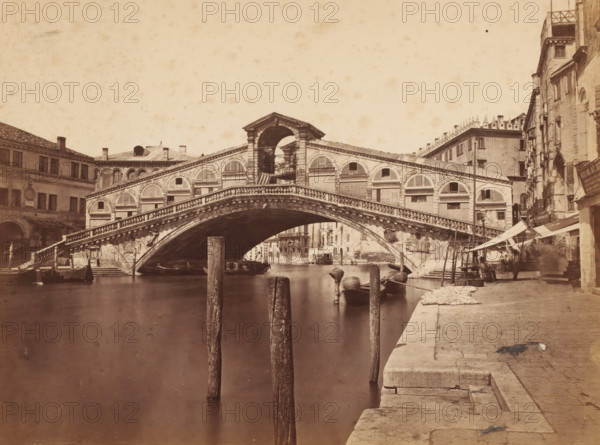 Ponte Rialto, Venice, View of the Rialto Bridge in Venice, Italy, photograph, 1872, photographer, pasteboard, paper, albumen print, albumendruk, height 187 mm x width 256 mm, bridge in city across river, canal, A stone arch bridge spans a calm canal, showing a central rounded opening beneath a balustraded walkway and decorative arched windows along its sides; wooden posts rise from the water in the foreground near small moored boats and gondolas; stone steps and a paved quay run along the right edge with awnings and stalls set up beside adjacent multi-story buildings that have shuttered windows and worn facades; on the left, more buildings with arched ground-level openings and windows frame the canal, and the sky above is pale and textured.