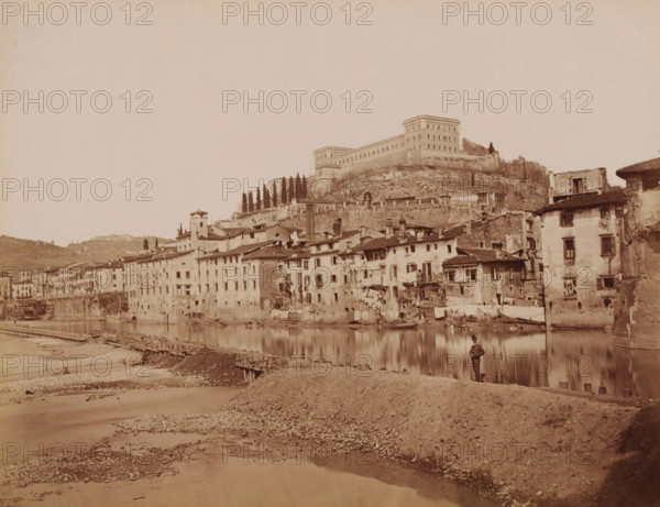 View of Verona and the Adige, Italy, photograph, inscription: 'Verona.', number: '13', number: '68', Verona, 1851 - 1900, photographer, pasteboard, paper, albumen print, albumendruk, height 304 mm x width 405 mm, city-view in general, 'veduta', river, castle, Adige, A riverside scene showing a row of multi-story stone and plaster buildings with tiled roofs lining the water's edge, many with small windows and balconies, some with laundry hanging outside, and small boats moored along the river; behind and above the buildings a steep rocky embankment rises to a large fortified structure with rectangular towers and rows of windows, flanked by a stand of tall narrow trees; in the foreground a gravelly riverbank slopes down to shallow water with a wooden pier or walkway extending along the shore, and a solitary figure standing near the water's edge.