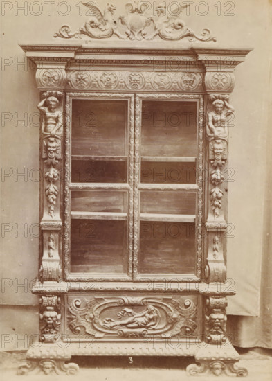 Walnut case, No 3 Mobile Noce, photograph, page, inscription: 'Ia. 320', inscription: 'Alto, 245. Largo, 152. Fondo, 36.', c. 1878 - c. 1885, photographer, paper, albumen print, height 135 mm x width 97 mm, Photo on unnumbered page, ornamental cupboard, An ornate wooden cabinet front with carved decorative elements including symmetrical columns framing a set of glass panels, sculpted human figures integrated into the vertical supports, carved floral and foliate motifs running along the frame, a horizontal frieze decorated with circular carved medallions, a carved crest with winged mythic creatures at the top, rectangular glass panes with visible shelves behind them, a carved lower panel featuring a reclining figure within an oval cartouche, stylized scrollwork and ornamental feet at the base, and a plain backdrop behind the piece.