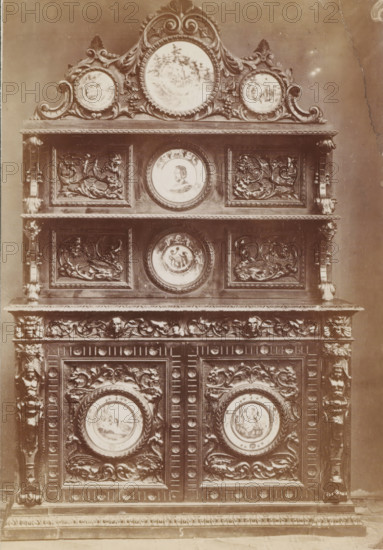 No 5 Buffet, Decorated sideboard, photograph, page, inscription: 'Ies. 350', inscription: 'Alto, 220. Largo, 155. Fondo, 60.', c. 1878 - c. 1885, photographer, paper, albumen print, height 136 mm x width 95 mm, Photo on unnumbered page, ornamental cupboard, A tall ornate wooden cabinet fills the frame, showing multiple carved panels, rounded medallions with painted scenes, and elaborate scrollwork; the top features a decorative crest with three circular painted plaques set within swirling carved foliage, beneath which are two horizontal shelves separated by carved pilasters, each shelf fronted by rectangular panels with raised relief floral and mythical motifs flanking central round painted portraits; lower sections continue the pattern with more carved panels and circular painted medallions encircled by twisted rope borders, all set within a richly carved frame that includes acanthus leaves, human masks, and ornamental borders.