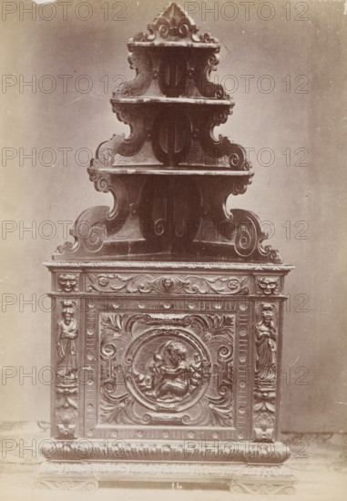 No 14 Cantoniera Noce, Walnut corner cabinet, photograph, page, inscription: 'Alta, 2m. Larga, 1m. Fonda, 68.', inscription: 'Ies 130.', c. 1878 - c. 1885, photographer, paper, albumen print, height 133 mm x width 95 mm, Photo on unnumbered page, ornamental cupboard, A tall ornate carved wooden structure with multiple tiered curved shelves rising to a pointed top, each shelf edged with scrolling motifs and decorative moldings, sits against a plain background. The lower section features a rectangular carved panel with an ornate circular medallion depicting two cherubic figures in bas relief, surrounded by acanthus leaves and scrollwork. Flanking the central panel are vertical pilasters carved with standing draped female figures and additional decorative motifs. The base rests on a molded plinth with carved foliate patterns and a textured surface. The image has a sepia tone and visible surface scratches and marks.