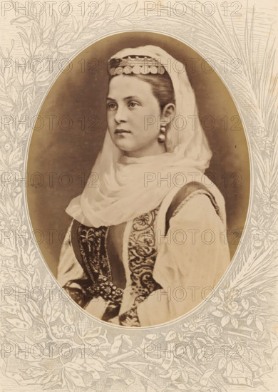 Olga, Queen of the Hellenes, Photographic reproduction of a painted portrait of Olga, Queen of Greece, photograph, reproduction, page, after painting by anonymous, c. 1875 - in or before 1880, photographer, paper, albumen print, height 189 mm x width 143 mm, Photo on unnumbered page., Person, historical persons - BB - woman, queen, empress, etc. (wife of a ruler), A sepia-toned oval portrait of a young woman shown from the chest up wearing a headscarf draped over her head and shoulders and a decorative cap with round ornaments across the front; she has smooth hair pulled back, a calm expression, and drop earrings. Her garment features ornate embroidered patterns on a dark central panel and lighter sleeves with visible trim. The oval portrait is set against a pale background with intricate floral and leaf line drawings framing the image.