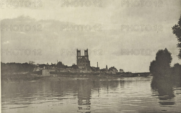 View of Mantes in the evening, Le Soir, à Mantes, photomechanical print, photographer: Achille Darnis, Limay, cliché maker: Fillon & Heuse, printer (person): Charles Wittmann, Paris, c. 1891 - in or before 1896, printer (person), paper, photoaquatint, height 129 mm x width 199 mm, city-view in general, 'veduta', river, dusk, evening twilight, Seine, Mantes-la-Jolie, A riverside scene in a muted monochrome palette shows a calm body of water in the foreground with gentle ripples reflecting dark vertical shapes. Across the water a low town silhouette stretches along the bank with clustered buildings and varied rooflines. A large structure with twin tower-like forms rises above the other buildings near the center, its vertical outlines mirrored faintly in the water. Trees and taller vegetation frame the right edge of the image as darker masses, and a broad sky occupies the upper portion with soft, diffuse cloud tones.