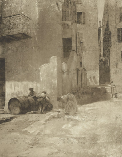 Street corner in Menton, Coin de rue, à Menton, photomechanical print, photographer: Robert Demachy, Menton, cliché maker: Fillon & Heuse, printer (person): Charles Wittmann, Paris, c. 1891 - in or before 1896, printer (person), Provence-Alpes-Côte d'Azur, paper, photoaquatint, height 214 mm x width 166 mm, street, A narrow, muted street scene rendered in sepia tones shows tall, weathered buildings flanking a rough, uneven roadway; a decorative iron balcony projects from the upper left wall, and several recessed windows and doorways punctuate the facades. In the foreground two figures lean over a large wooden barrel lying on its side while another figure bends forward near a small bucket; farther down the street, a solitary figure stands near a corner. The ground is textured with patches of light and shadow, and the overall surface of the image has a grainy, worn appearance.