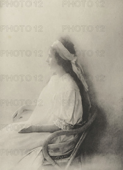Portrait, Portrait of an unknown woman, photomechanical print, photographer: baron O. de Laudon, cliché maker: Fillon & Heuse, printer (person): Charles Wittmann, Paris, c. 1891 - in or before 1896, printer (person), paper, photoaquatint, height 184 mm x width 137 mm, historical persons not known by name - BB - woman, A sepia-toned portrait of a young woman in profile seated on a wicker chair, wearing a light, loose dress with puffed sleeves and lace trim at the cuff, her long dark hair tied back with a wide light-colored ribbon that drapes down behind her, her hands resting gently in her lap, and a softly lit plain background.