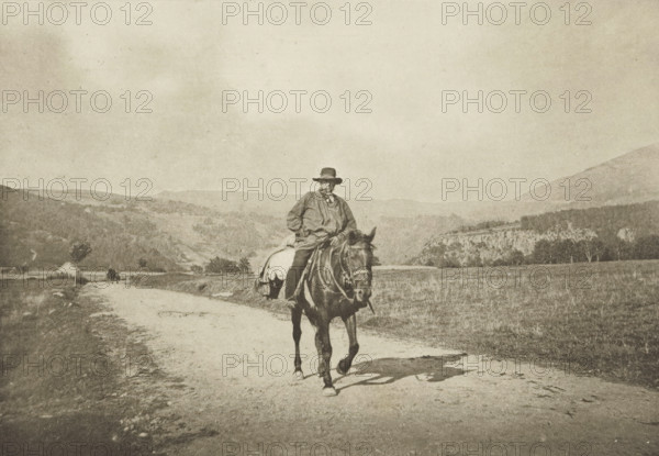 Retour du Moulin (Mont-Dore), Rider on a country road, Mont-Dore, photomechanical print, photographer: G. de Magnitot, cliché maker: Fillon & Heuse, printer (person): Charles Wittmann, Paris, c. 1891 - in or before 1896, printer (person), paper, photoaquatint, height 129 mm x width 185 mm, riding a horse, ass, or mule, rider, horseman, (high) hill, A sepia-toned scene shows a person wearing a wide-brimmed hat and loose outer garment seated on a saddled horse that is walking along a dirt road, with the rider holding the reins and looking slightly to the side; the road stretches into a pastoral landscape with grassy fields and low hills, scattered trees, and a small building near the roadway in the distance, while further hills and a cliff-like formation rise beyond and the sky above is pale and expansive.