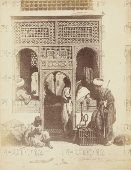 Photo reproduction of the painting 'A barber's shop in Cairo' by Willem de Famars Testas, photograph, reproduction, page, photographer: Binger & Chits, after painting by Willem de Famars Testas, c. 1866 - in or before 1871, Binger & Chits, Willem de Famars Testas, photographer, after painting by, paper, albumen print, height 190 mm x width 147 mm, barber, barbershop, A monochrome scene shows an ornate doorway with carved and latticed panels and arched openings; patterned screens fill the upper portion and decorative motifs frame the central archway. Several figures in flowing robes and head coverings stand and sit around the entrance, one seated on the ground with folded legs, another leaning near a window opening, and others clustered in and near the doorway. A tall figure holds a long staff and a coiled hose or pipe rests on a wicker crate beside them. Rounded sacks or bundles lie near the walls and a small bird is visible on the ground in front of the entrance.
