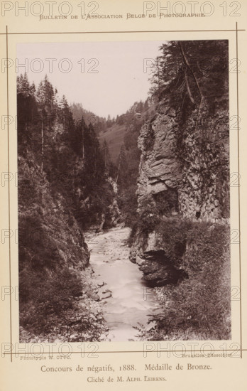 Rock formations in the Tamina valley, Switzerland, page, photomechanical print, inscription: 'Concours de négatifs, 1888. Médaille de Bronze.', photographer: Alphonse Leirens, Tamina Gorge, cliché maker: Wilhelm Otto, Brussels, 1886-09-07, printer (person), Taminakloof, pasteboard, paper, collotype, photoprint, height 170 mm x width 120 mm, Print on unnumbered page., brook, rock-formations, Tamina, A narrow river flowing between steep rocky cliffs and wooded slopes, with trees lining the ridges and growing on the cliff faces, a rocky overhang on the right side, vegetation and shrubs along the riverbanks, a distant hillside with scattered trees, and the photograph printed on a bordered page with a printed heading at the top and printed captions along the bottom.