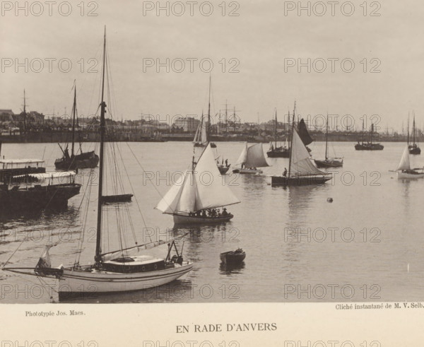 Sailing ships at the port of Antwerp, En rade d'Anvers, page, photomechanical print, inscription: 'Section d'Anvers - Exposition de 1889', photographer: Victor Selb, Antwerp, cliché maker: Joseph Maes, c. 1883 - in or before 1888, printer (person), paper, collotype, photoprint, height 115 mm x width 156 mm, Print on unnumbered page., harbour, sailing-ship, sailing-boat, A waterfront scene showing multiple sailboats and small boats on calm water with visible masts, rigging, and unfurled sails; a larger sailboat in the foreground with a cabin and visible deck fittings, smaller sailboats with triangular sails scattered across the water, and a pair of rowboats; reflections of the boats on the water surface; a distant shoreline with buildings and additional masts lining the horizon; a mostly overcast sky above.