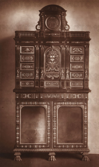 Cabinet in the style of Louis XIII, from the collection of Christopher Beckett Denison, page, photomechanical print, number: '1868', c. 1880 - in or before 1885, paper, Woodburytype, height 195 mm x width 121 mm, Print on third unnumbered page after page 178., ornamental cupboard (wood and other phytogenic material), A tall ornate wooden cabinet photographed against a plain backdrop, featuring multiple stacked sections with carved moldings and decorative inlays; the uppermost portion has a rounded pediment with a circular carved medallion flanked by scroll-like ornaments and small balustrades, below which are rectangular panels with intricate scrollwork and floral motifs; the central section displays a recessed arched panel filled with elaborate symmetrical foliage and foliate patterns surrounded by fluted pilasters and framed drawers and panels bearing carved ornament; the lower section includes two large recessed door openings bordered by patterned inlay and narrow vertical columns o...