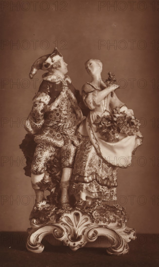 Two porcelain statues, depicting summer and winter, page, photomechanical print, number: '3101', after sculpture by anonymous, c. 1880 - in or before 1885, after sculpture by, paper, Woodburytype, height 194 mm x width 121 mm, summer, 'Aestas', 'Estate' (Ripa), winter, 'Hyems', 'Inverno' (Ripa), piece of sculpture, reproduction of a piece of sculpture (stone, arts), A sepia-toned photograph of an ornate porcelain figurine depicting a dressed man and woman standing side by side on an elaborate scrollwork base. The man wears a patterned coat, breeches, and a hat with a long, drooping tip, his attire showing intricate floral and geometric designs and textured furlike trim. The woman wears a patterned dress with ruffles and a decorative apron filled with sculpted flowers, and she holds a small bouquet near her shoulder while her head tilts slightly toward the man. Both figures have detailed facial features and stylized hair, and the entire scene rests on a richly decorated base with carved...