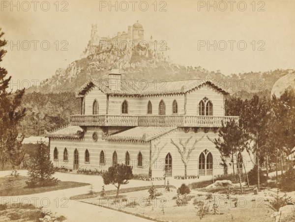 Swiss chalet of the Countess of Edla in Sintra, Swiss chalet in New Garden (Palace of the Pena in the distance), photograph, page, Sintra, c. 1865 - in or before 1870, photographer, paper, albumen print, height 147 mm x width 198 mm, Photo on second unnumbered page after page 4., farm or solitary house in landscape, palace, Palácio da Pena, A sepia-toned scene shows a large wooden house with decorative trim along the roofline and pointed arched windows, including a pair of tall arched windows on one side and several smaller arched windows along the lower level; the house has a balcony with balustrade, a chimney, and patterned roof tiles. In front of the house are small planted shrubs, a pathway, and a few young trees. Behind the house a densely wooded area rises into rocky slopes, and atop a rocky hill a stone fortress-like structure with towers and a domed section is visible against a pale sky.