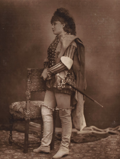 Portrait of Marguerite Ugalde, page, photomechanical print, photographer: Félix Nadar, cliché maker: Lemercier, c. 1880 - c. 1885, paper, Woodburytype, Woodburytypie, height 163 mm x width 201 mm, Person, Marguerite Ugalde, historical persons - BB - woman, portrait of actor, actress, opera-singer (female), A sepia portrait of a person standing in profile wearing a patterned, fitted garment with decorative buttons and striped sleeves, a draped cloak over one shoulder, and thigh-high boots with ruffled garters, holding a sheathed sword with an ornate hilt and resting one hand on the back of an upholstered chair with fringe and carved wooden legs; the background is plain and the floor covered with a patterned rug and a draped fabric.