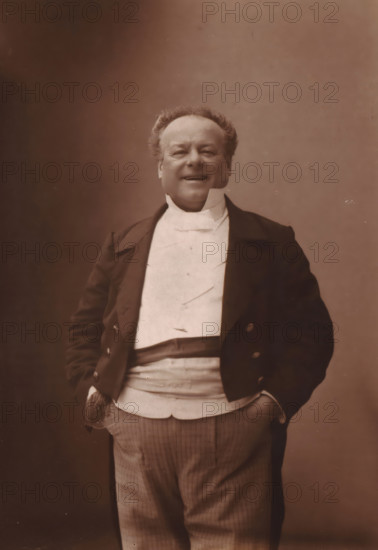 Portrait of Jean Berthelier, page, photomechanical print, photographer: Félix Nadar, cliché maker: Lemercier, c. 1880 - c. 1885, paper, Woodburytype, Woodburytypepie, height 242 mm x width 173 mm, Person, Jean-François Berthelier, historical persons, portrait of actor, actress, A man standing in a formal dark jacket with a light waistcoat and a high stiff collar, his hands tucked into his pockets, smiling with a round face and short curly hair, wearing a dark sash around his midsection and patterned trousers, photographed against a plain neutral background.