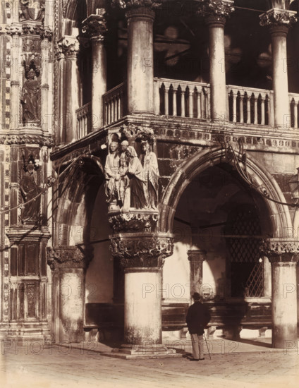 Palazzo Ducale, photograph, c. 1850 - c. 1870, photographer, albumen print, height 583 mm x width 446 mm, height 346 mm x width 267 mm, Venice, Doge's Palace, A sepia-toned photograph of an ornate stone façade featuring tall columns, arched openings, carved capitals, and balustrades, with sculpted figures in relief mounted on a projecting bracket above an arch; a man wearing a dark jacket and light trousers stands with a cane at the base of a central column, casting a shadow on the wall behind him; decorative vertical panels with recessed niches and statues flank the left edge of the image; wrought metal brackets curve above the arches and a barred window is visible through one arch; the ground is paved and shows shadows from the architectural elements.