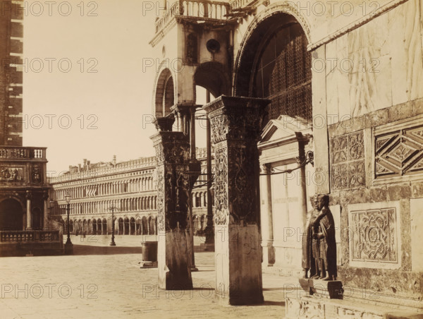 Piazza S. Marco presa dalla Porta della Carta. Venezia., photograph, photographer: Carlo Naya, 1875, albumen print, albumendruk, height 271 mm x width 353 mm, height 318 mm x width 455 mm, Venice, A sepia-toned architectural scene shows a row of ornate stone columns and arches with carved capitals and decorative reliefs, a tall building facade with repeated arched windows stretching into the distance, a paved open plaza in the foreground, a metal lamppost standing on the plaza, a sculpted standing figure in armor positioned against a textured stone wall with carved panels, a small triangular pediment above a recessed doorway, patterned metal grilles set into one of the arches, and a balustraded balcony visible at the left edge.