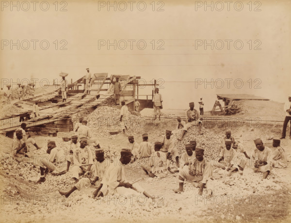 United States, prisoners at Rock Fort Kingston (former title), Prisoners at Rockfort, Kingston, Jamaica, photograph, c. 1860 - c. 1890, photographer, albumen print, height 301 mm x width 396 mm, height 261 mm x width 338 mm, Rockfort, A wide sepia photograph shows a shoreline worksite with many men wearing light-colored loose clothing and caps seated on and standing among large piles of small rocks and gravel; several men hold hand tools while others sit on the gravel piles or rest on wooden planks. In the midground there are wooden ramps and sluice-like structures with slatted screens and chutes leading down to the piles, and a few men stand on and beside these structures, one carrying a rectangular tray on his head. Narrow rails and wooden platforms run across the site toward the water, and small wooden sheds and workbenches are visible near the right edge. The calm water fills the background horizon, and the sky above is pale and mostly empty. The image has an overall aged, grainy, ...