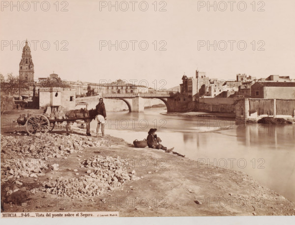 Murcia-Vista del puente sobre el Segura, photograph, photographer: Juan Laurent, c. 1857 - c. 1880, albumen print, height 250 mm x width 338 mm, A wide riverside scene with a stone bridge spanning the water and buildings lining the far bank, a tall bell tower visible on the left, masonry walls and varied rooftops across the background, and a calm reflective river in the foreground. On the near bank there is a dirt and rocky shoreline, a man standing beside a cart hitched to a donkey, a seated figure on the ground near a basket, scattered stones and rubble, and small structures and walls close to the bridge.