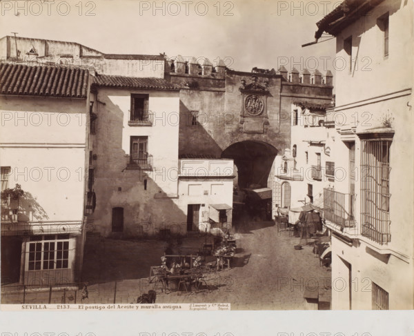 Sevilla-El postigo del aceite y muralla antigua, photograph, photographer: Juan Laurent, c. 1857 - c. 1880, albumen print, height 253 mm x width 337 mm, Andalusia, Seville, A sunlit street scene framed by whitewashed buildings with tiled roofs and iron balconies, showing a cobbled roadway leading toward a large arched gateway set into an old stone wall topped with crenellations, the gateway flanked by a decorative circular relief panel and sculpted details above it, market stalls and wooden tables displaying goods arranged along the street beneath awnings and cloth coverings, shadows cast across façades and the pavement, narrow windows and doorways punctuating the building faces, and various small architectural elements like cornices, drainpipes, and a barred ground-floor opening.