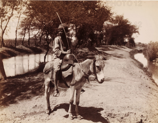 Unknown boy on a donkey, Ane de Place, photograph, number: '148', photographer: Jean Pascal Sébah, Middle East, 1888 - 1900, paper, albumen print, height 210 mm x width 270 mm, boy (child between toddler and youth), ass, donkey, A sepia-toned photograph shows a person sitting on a saddled donkey on a dirt path beside a narrow water canal, the person wearing a headwrap and loose clothing and holding a long stick upright, the donkey facing slightly toward the camera with a bridle and visible saddlebag, trees with leafy branches line the canal and cast shadows across the path and water, and the scene extends into a distant shaded tree-lined background under a bright sky.
