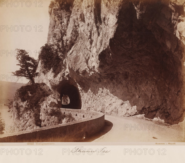 View of the Merligenstrasse along the Thunersee, Switzerland, Merligenstrasse, photograph, number: '1603', inscription: 'Thuner See', number: '13932', photographer: Giorgio Sommer, Thunersee, 1857 - 1914, pasteboard, paper, albumen print, height 334 mm x width 450 mm, public road, mountains, A winding road curves along a rock face with a low stone barrier following the curve, leading into a dark arched tunnel carved into the cliff; rough textured rock dominates the right side of the scene while sparse vegetation and a few small trees cling to ledges on the left, and the foreground shows the dirt surface of the road with a shadow cast along the inner curve.