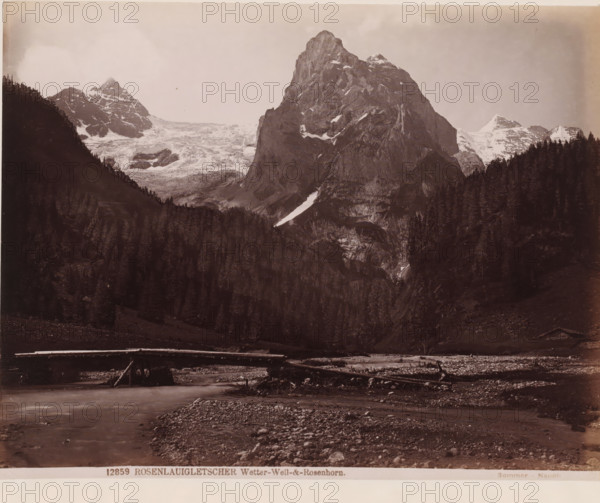 View of the Rosenhorn, Switzerland, Rosenlaigletscher. Wetter-Well und Rosenhorn, photograph, inscription: '1665', inscription: '150=', number: '12859', number: '140', photographer: Giorgio Sommer, Berner Alpen, 1857 - 1914, Alpen, pasteboard, paper, albumen print, albumendruk, height 333 mm x width 435 mm, brook, dale, valley, mountain-top, peak, Alps, Bernese Alps, Rosenhorn, A wide sepia-toned landscape photograph showing a steep rocky mountain with patches of snow rising behind a dense dark forest of tall conifer trees, flanked by additional snow-capped peaks and a glacier visible on one side, with a shallow river or stream in the foreground flowing past a rocky, pebble-strewn bank and a simple log bridge spanning the water.