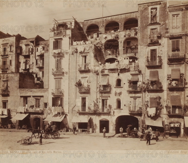 Facades with drying laundry on Via Nuova Marina in Naples, Italy, Vecchie case in Via della Marina, Napoli, photograph, inscription: '100.=', number: '1599', number: '137', number: '12435.', photographer: Carlo Brogi (possibly), Naples, publisher: Edizioni Brogi, Florence, 1881 - 1900, Tuscany, pasteboard, paper, albumen print, height 316 mm x width 448 mm, hanging the wash to dry, facade (of house or building), Via Nuova Marina, A row of weathered multi-story buildings with balconies and open archways fills the scene, their facades showing peeling plaster and patches of exposed brick; laundry hangs from numerous balconies and lines strung across the fronts, billowing and draped over railings; some balconies contain plants in pots and small railings, while others show window shutters in various states of openness or closure; beneath the buildings are shaded shopfronts and arch entrances, with a few people standing or sitting near the doorways and under awnings; in the open paved area i...