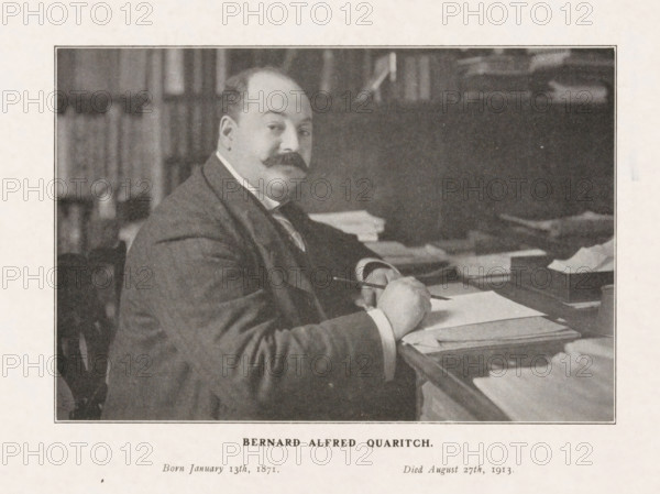 Bernard Alfred Quaritch, Portrait of bookseller Bernard Alfred Quaritch behind his desk, print, photograph, after 1913-08-27, printmaker, photographer, paper, printing, height 225 mm x width 287 mm, Person, book-shop, bookseller, A sepia-toned photograph of a seated man with a rounded face and a prominent mustache, wearing a dark suit and a white shirt collar, shown from the waist up at a desk covered with stacks of papers and folders; his right hand holds a pen above an open sheet while his left rests on the desk, and behind him are blurred bookshelves and indistinct objects on the surface behind the desk.