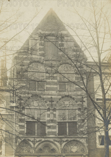 Facade of a house on the Koornmarkt in Delft, Koornmarkt WZ No. 81. Early Renaissance facade, photograph, number: '27', inscription: 'Delft. 1st half of the 16th century.', inscription: 'The right of reproduction of these photos is reserved by the Publisher.', Delft, c. 1900 - c. 1904, photographer, paper, pasteboard, gelatin silver print, height 167 mm x width 120 mm, urban housing, ornament derived from architectural details, Koornmarkt, A tall narrow building facade with a steep triangular roof, masonry bands visible across the exterior, multiple rectangular windows with some shutters closed, ornate arched decorative reliefs above lower windows, bare tree branches partially obscuring the view, adjacent building walls visible at the sides, and a muted, aged photographic tone.