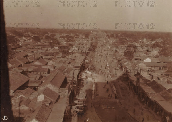 Bird's-eye view of a long shopping street in a vast city, China, photograph, inscription: 'Douairière Hoeufft-van de Spie etc. wedding announcement', photographer: attributed to Jan Adriani, 1907, Jan Adriani, photographer, Attributed to, paper, daylight collodion silver print, daylight collodion silver print on photo paper, height 80 mm x width 105 mm, city-view in general, 'veduta', street, A sepia-toned aerial view of a wide street running through a dense area of low buildings with tiled roofs, the street lined with small structures and stalls and dotted with many tiny figures walking or standing, trees scattered among the rooftops, a distant hazy horizon, and a vertical dark strip along the left edge of the image.
