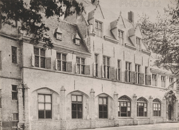 View of the Abbey of Middelburg from the inner courtyard, Former residence of the abbot, Ancien logement de l'abbé, Views of the Abbey in Middelburg, Abbey in Middelburg, Abbaye de Middelbourg, photomechanical print, cliché maker: Straaten, Gebroeders van, Middelburg, in or after 1900, Straaten, photographer, cliché maker, paper, collotype, photogravure, height 150 mm x width 202 mm, abbey, monastery, convent, Roman Catholic church, Abbey of Middelburg, A stone building facade with arched ground-level windows and rectangular upper windows, some of which have open wooden shutters; a steep tiled roof with small dormer windows and larger gabled dormers above; decorative molding and stone trim around windows and along the roofline; leafy tree branches partially framing the top corners of the image; a paved area in front of the building.