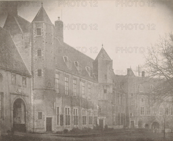 View of the State Archives buildings in the Abbey in Middelburg, The State Archives buildings on the north side of the large courtyard in 1888, The buildings of the State Archives, Views of the Abbey in Middelburg, Abbey in Middelburg, Abbaye de Middelbourg, photomechanical print, cliché maker: Straaten, Gebroeders van, Middelburg, in or after 1900, photographer, paper, collotype, photogravure, height 150 mm x width 186 mm, courtyard, abbey, monastery, convent, Roman Catholic church, archive, Abbey of Middelburg, A sepia-toned photograph of an old stone building with steep pitched roofs and multiple towers, showing rows of tall windows along the facade and several dormer windows set into the roof; a large arched doorway is visible at one end and smaller doorways and window openings punctuate the ground level; a bare-branched tree stands on the right side of the scene, its limbs extending into the sky above the roofline; portions of the street and pavement run in front of the building.