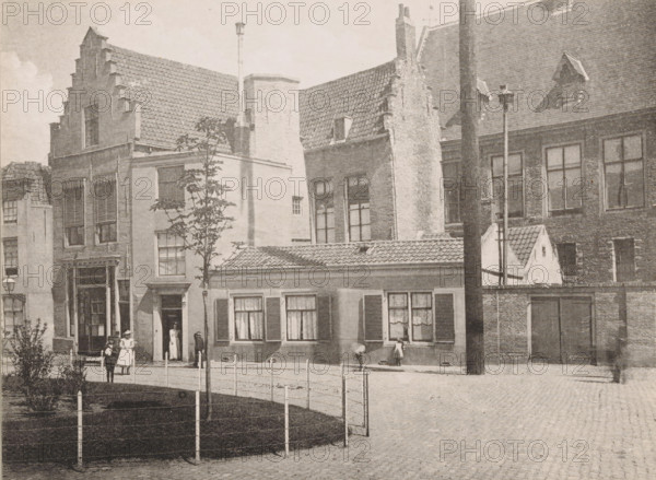 House on the Balance, purchased for the Provincial Water Authority, before the restoration, 1895, Maison sur la place de la Balance, View of the House on the Balance near the Abbey of Middelburg, Views of the Abbey in Middelburg, Abbey in Middelburg, Abbaye de Middelbourg, photomechanical print, cliché maker: Straaten, Gebroeders van, Middelburg, in or after 1900, photographer, paper, collotype, photogravure, height 150 mm x width 201 mm, abbey, monastery, convent, Roman Catholic church, Abbey of Middelburg, A row of brick and stucco buildings with tiled roofs and stepped gables lines a cobblestone street, several windows with shutters and lace curtains visible on the lower facades and tall multi-pane windows on the upper floors, a narrow doorway with a person standing inside and a few people—some wearing dresses and hats—standing and walking on the sidewalk, a small fenced grassy area with a young tree in front, a low building with shuttered windows and a tiled roof at street level, a...