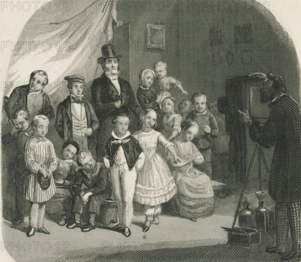 Family posing for a group photo, print, album page, Print is part of an album. Print is loose in the album next to the design drawing., printmaker: Johann Wilhelm Kaiser (I) (possibly), 1840 - 1900, paper, etching, height 126 mm x width 138 mm, family group, especially parents with their child(ren), taking photographs of model, sitter, camera, photography, A monochrome engraving showing a group of people gathered in an interior space, including men, women, and children in period clothing; a tall man wearing a top hat and formal coat stands near the center with a hand tucked to his chest, while a woman and a child stand close beside him and another woman sits holding fabric in her lap; several boys and girls in dresses and jackets occupy the foreground, some standing and some seated, with varied facial expressions and postures; on the right edge a bearded man in a dark robe faces the group and gestures with one hand, and near him on the floor are glass bottles and a small box with a lon...