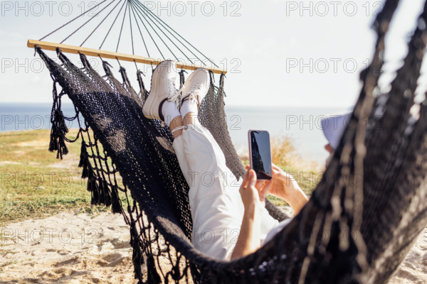 A young woman in casual clothes holds a phone in her hands and lies in a hammock. A girl in a summer hat uses a mobile g
