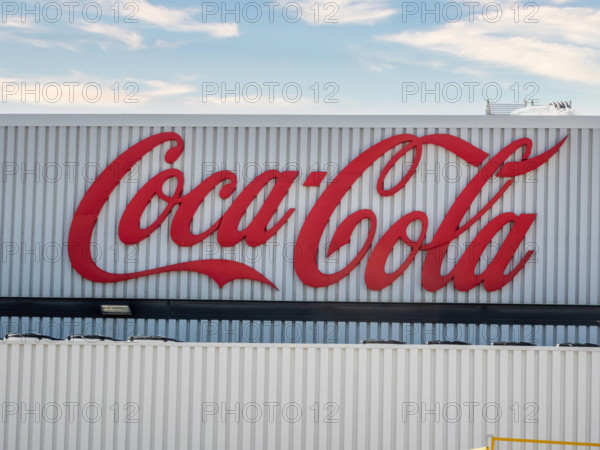 Ein großes, rotes Coca-Cola-Logo prangt auf einem weißen Gebäude mit vertikalen Streifen unter einem teilweise bewölkten Himmel.